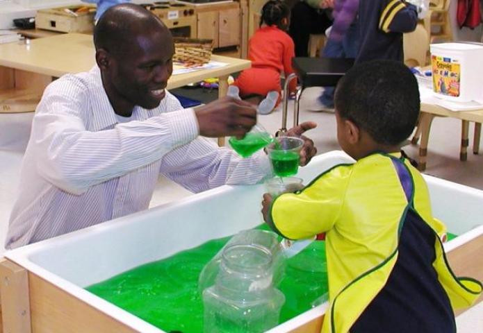 Child with a teacher filling cups with green liquid.