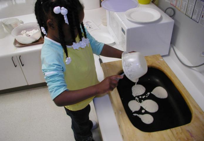 Young girl pouring pancake batter into a griddle.