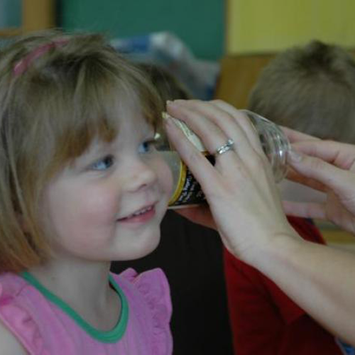 young girl listening to a glass jar while teacher plucking a rubber band